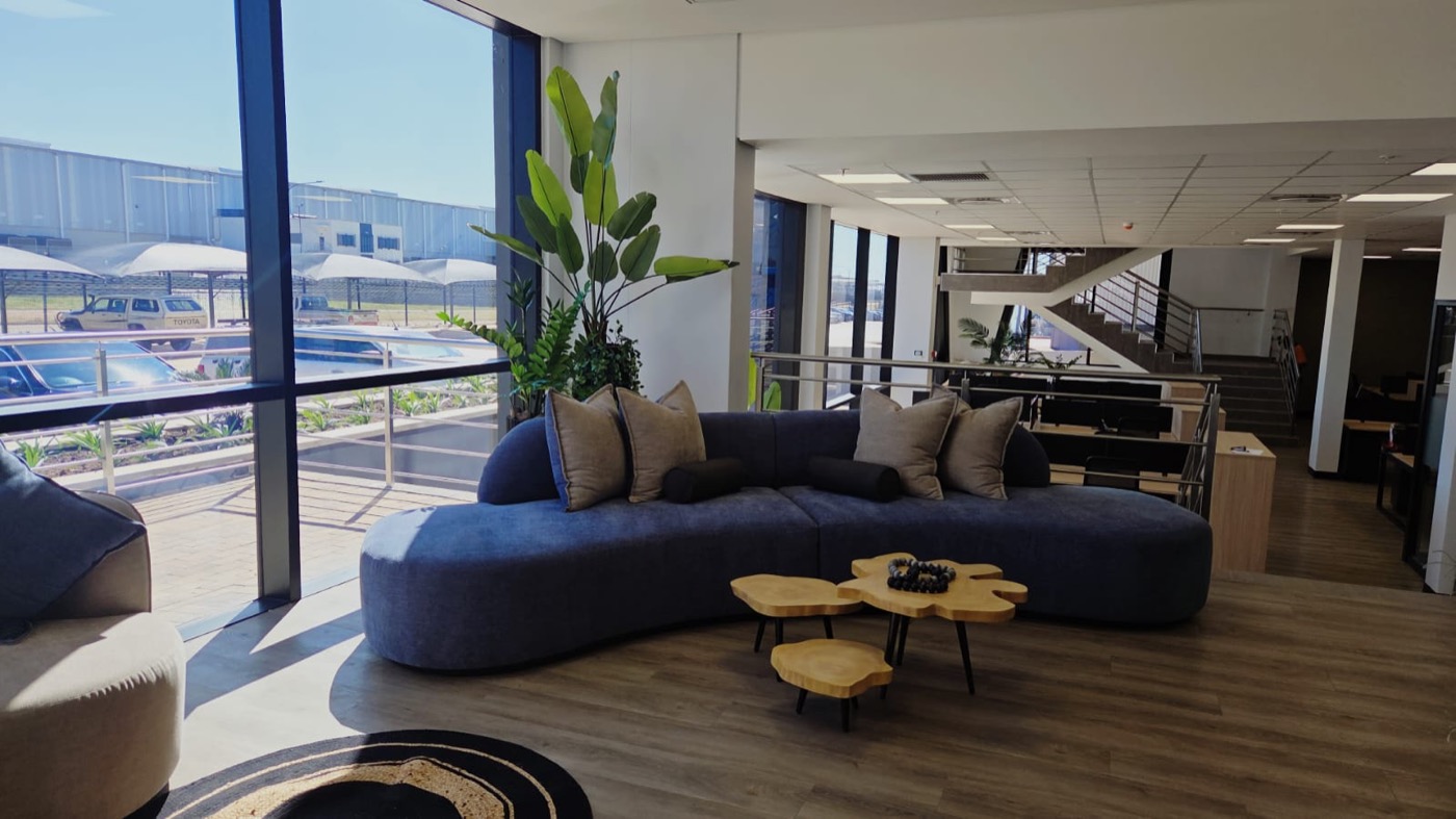 Completed open lounge space with large windows, curved sofa, plant styling, and warm timber tables
